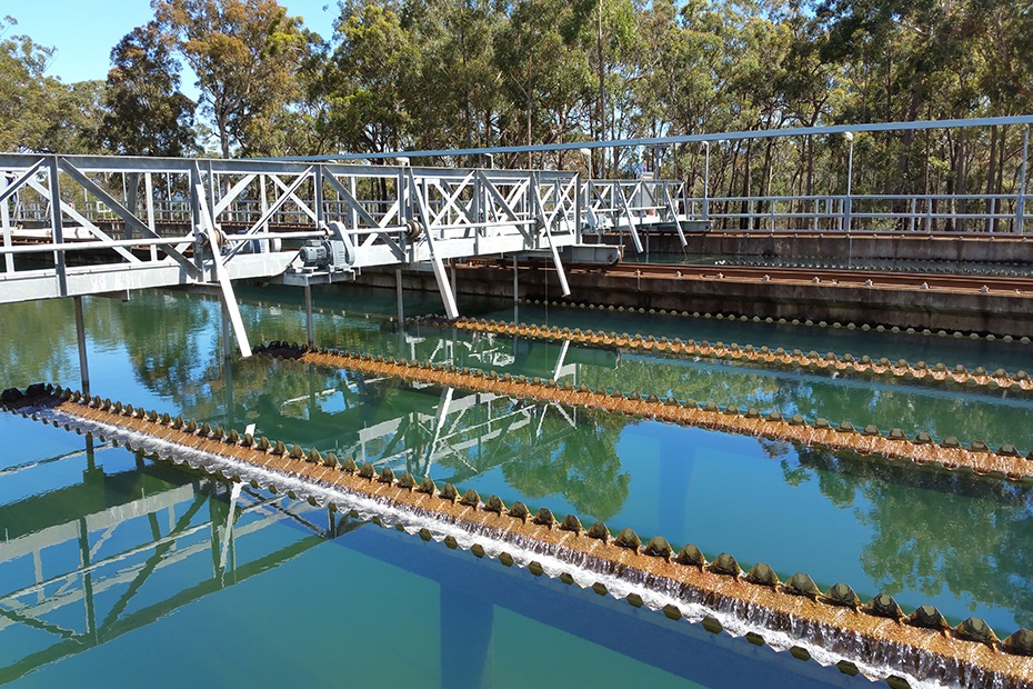 A picture of a water treatment plant. There is a large pool of still water with big metal pipes on the surface, and a metal catwalk over it with trees in the background.